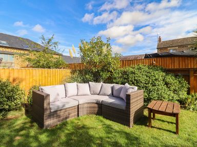 An outdoor patio area with a curved rattan sofa and cushions next to a wooden table in a garden with bushes and a wooden fence at The Stables in Great Tew near Chipping Norton