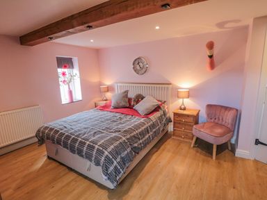 A bedroom with a bed two bedside tables with lamps a pink chair a wall clock and flowers on a windowsill at Ericas eaven in Farndale near Kirkbymoorside
