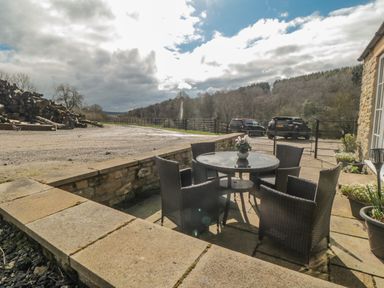 A patio with a round glass table and four wicker chairs near a stone building and parked cars in a rural setting at Ericas eaven in Farndale near Kirkbymoorside