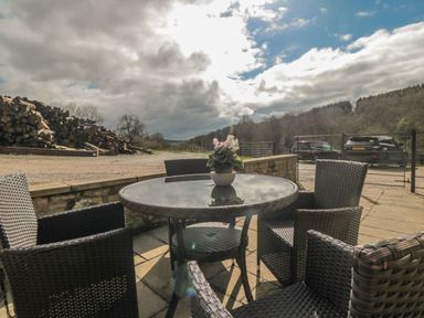 An outdoor patio with a round glass table and four wicker chairs a small potted plant on the table logs stacked in the background and two parked cars near a forested area at Ericas eaven Farndale near Kirkbymoorside