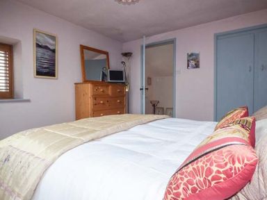 A bedroom with a bed dressed in white and beige bedding a wooden chest of drawers with a mirror small TV and a lamp with blue framed doors and a window with wooden shutters at High House Newton in Cartmel near Cartmel