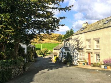A street with houses on the right and trees on the left with hills and a cloudy sky in the background at High House Newton in Cartmel near Cartmel