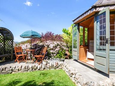 A garden patio with wooden chairs a table and umbrella next to a small garden house with open doors at High House in Newton in Cartmel near Cartmel