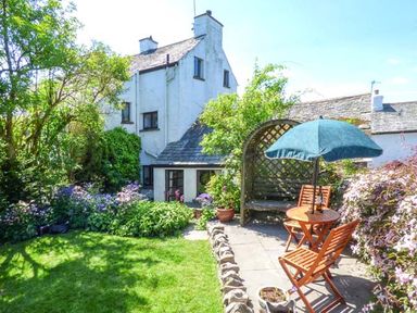 A garden with green grass bushes and flowers a wooden table with chairs and a blue umbrella at High House in Newton in Cartmel near Cartmel
