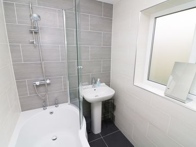 A bathroom with a bathtub shower combination a pedestal sink a small mirror on the windowsill and tiled walls at Wysteria Cottage in Windermere