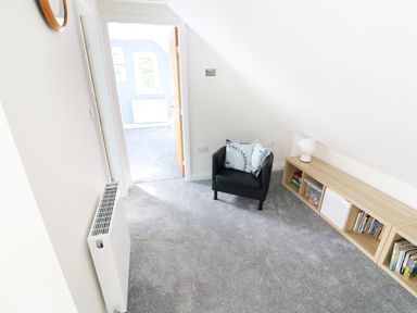 A room with grey carpet a black armchair with pillows a wooden shelf with books and a lamp at Wysteria Cottage in Windermere