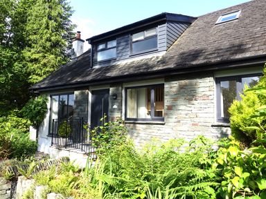 Exterior view of a stone and wood house with front door windows and a garden with various plants and shrubs at Wysteria Cottage in Windermere
