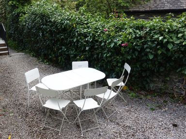 An outdoor seating area with a white table and six white chairs on gravel next to a green hedge at Wysteria Cottage in Windermere