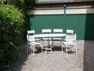 An outdoor seating area with a white table and six white chairs on a gravel surface next to a green fence and plants at Wysteria Cottage in Windermere