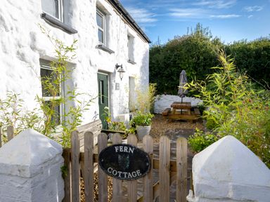 An outdoor area with a wooden gate labeled Fern Cottage white cottage walls green door wooden bench potted plants and a wooden picnic table with a closed umbrella in Aberporth