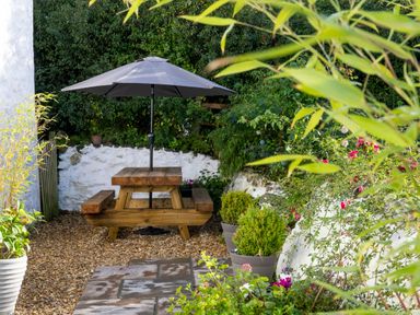 An outdoor area with a wooden picnic table and gray umbrella surrounded by plants and white stone walls at Fern Cottage in Aberporth