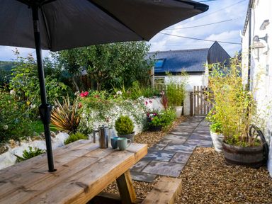 A garden patio with a wooden table and umbrella surrounded by plants and a stone path at Fern Cottage in Aberporth