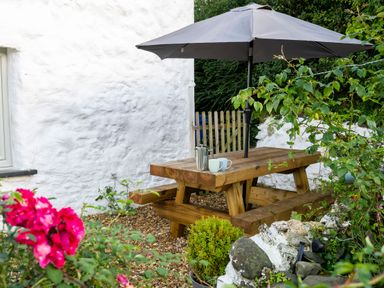 An outdoor wooden picnic table with a large umbrella and coffee items next to a white wall and garden at Fern Cottage in Aberporth