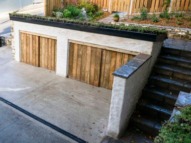 A double wooden garage door with stone walls and concrete driveway next to stone steps with plants above at Fern Cottage in Aberporth