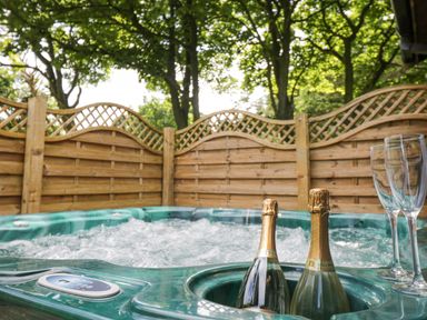 A bubbling outdoor hot tub with two bottles of champagne and two empty glasses in a wooden fenced area with trees at Pond Farm Woodlands in Ugthorpe