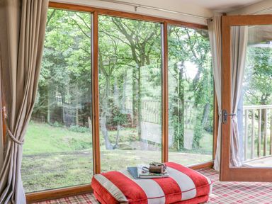 A room with a large glass door and windows looking out to trees and grass with a red and white striped cushion on a plaid carpet at Pond Farm Woodlands in Ugthorpe