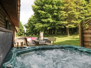 An outdoor hot tub with bubbling water next to a wooden table and benches surrounded by trees at Pond Farm Woodlands in Ugthorpe