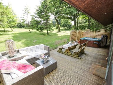 An outdoor patio with wooden seating a hot tub and a lawn with trees at Pond Farm Woodlands in Ugthorpe