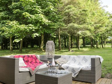An outdoor seating area with wicker sofas cushions blankets drinks and glassware in a wooded garden at Pond Farm Woodlands Ugthorpe