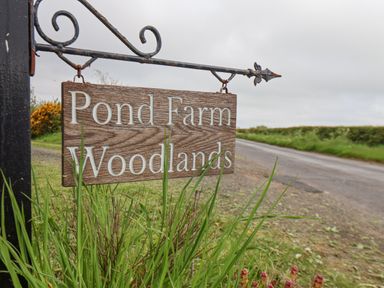 A wooden sign reading Pond Farm Woodlands hanging from a black metal bracket next to a road at Pond Farm Woodlands Ugthorpe