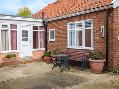 An outdoor patio area with a wooden bench and metal table next to a brick house with white windows and flower pots at Glenhaven in Sweffling near Snape