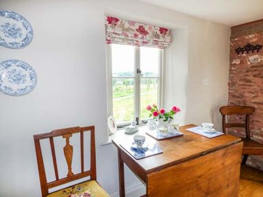 A small wooden dining table with two chairs and teacups near a window with floral curtains at Rose Cottage in Welsh Newton