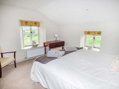 A bedroom with a bed a wooden chair a small dressing table and two windows with yellow floral blinds at Rose Cottage in Welsh Newton