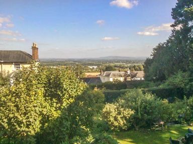 A garden with trees and shrubs and houses in the distance with a view of the countryside at Woodland Cottage in Malvern