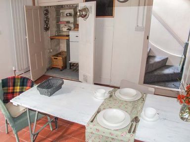 A dining table set for two with white dishes and cups a basket on a chair and a small pantry with pots and shelves at Woodland Cottage in Malvern