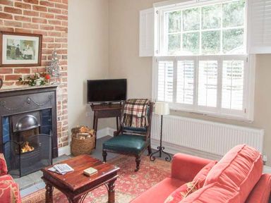 A living room with a fireplace a red sofa a wooden coffee table a chair with a blanket a television and a window with shutters at Woodland Cottage in Malvern