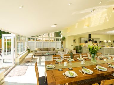 A dining area with a large wooden table set for a meal and an adjoining living room with sofas and a kitchen in the background at Old Rectory House in Caersws