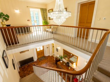 A staircase with wooden handrail leading to an upper landing with furniture and a chandelier at Old Rectory House in Caersws