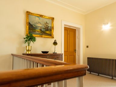 A hallway with a wooden banister chest of drawers with a plant lamp and bowl a radiator and a wooden door at Old Rectory House in Caersws