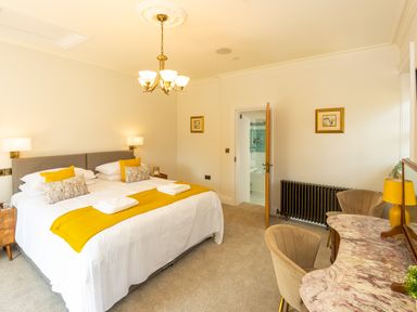 A bedroom with a double bed yellow cushions side tables a chandelier a dressing table with chairs and a door leading to a bathroom at Old Rectory House in Caersws