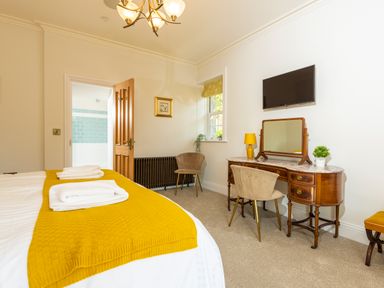 A bedroom with a bed covered with a yellow blanket towels on the bed a wooden door a vanity table with a mirror two chairs a lamp a wall mounted tv and a window at Old Rectory House in Caersws