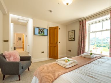 A bedroom with a bed, gray armchair with a pink cushion, wall-mounted TV, window with curtains, and an open wooden door at Old Rectory House in Caersws