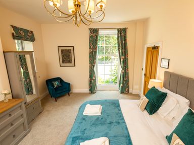 A bedroom with a bed green pillows and blanket a wooden dresser a green armchair floral curtains and a chandelier at Old Rectory House in Caersws