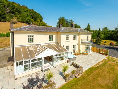 A large cream house with a glass conservatory a patio with outdoor seating a hot tub and surrounding trees at Old Rectory House in Caersws