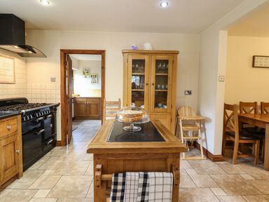 A kitchen with wooden cabinets a black stove a wooden island with a cake under a glass dome a wooden cupboard and a dining area with wooden chairs at Ty Newydd in Llangadfan