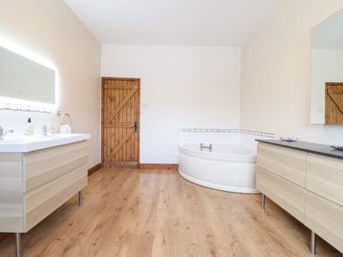 A bathroom with wooden floor a wooden door a corner bathtub double sink and two large mirrors at Ty Newydd in Llangadfan