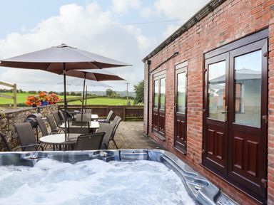 An outdoor patio with a hot tub tables and chairs with umbrellas and a brick building with wooden doors at Ty Newydd in Llangadfan