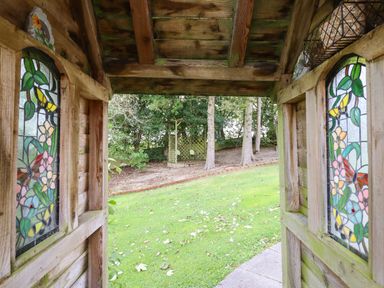 View from a wooden structure with stained glass windows to a garden with grass trees and a wooden seating area at Ty Newydd in Llangadfan