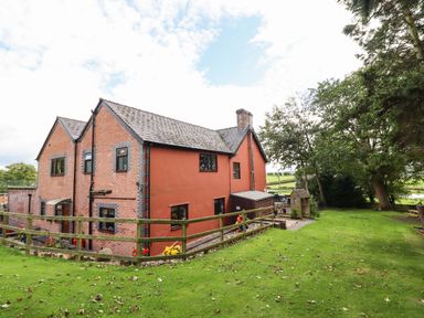 A brick and red plaster house with a fenced garden and green grass surrounded by trees at Ty Newydd in Llangadfan