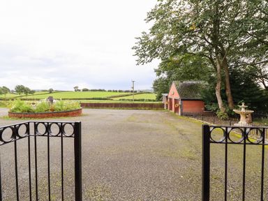 An open driveway with a black metal gate a brick round flower bed trees and a small red brick building at Ty Newydd in Llangadfan