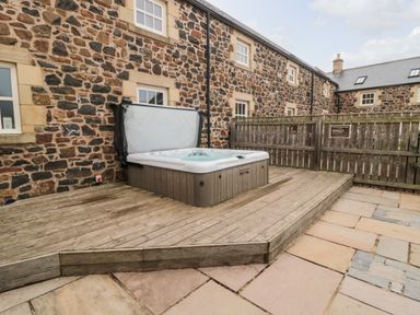 An outdoor patio area with a hot tub on a wooden deck next to a stone building at High Hemmel House in Embleton