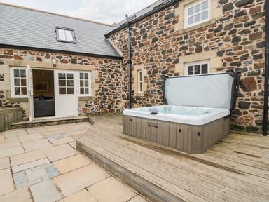 An outdoor patio area with a hot tub on a wooden deck next to a stone building at High Hemmel House in Embleton