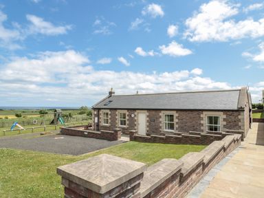 A brick house with a paved walkway and fenced grass area with playground equipment at Home Stead Cottage in Embleton
