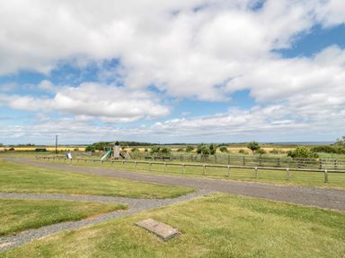 A playground with a slide and swings on grassy fields with a gravel path and a fence in a rural area at Home Stead Cottage in Embleton