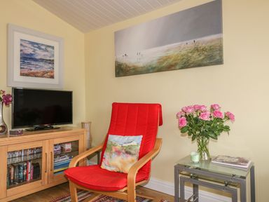 A sitting area with a red cushioned chair a wooden TV stand with a television and a side table with pink flowers at Tiggy Winkle in St Merryn near Padstow
