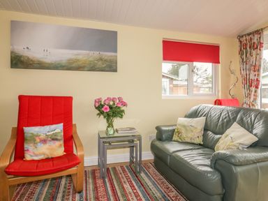A living room with a red armchair and a leather sofa with cushions and a side table with flowers at Tiggy Winkle in St Merryn near Padstow